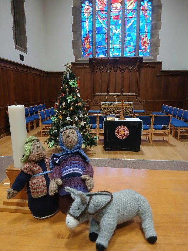 Knitted figures resembling Mary and Joseph, a stuffed donkey, a decorated Christmas tree, and a large candle inside a church with a stained glass window in the background.