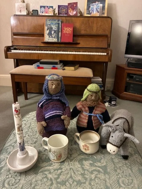 Knitted stuffed animals, including two people, a donkey, and a candle, arranged on a patterned tablecloth in front of a wooden piano with books on top.