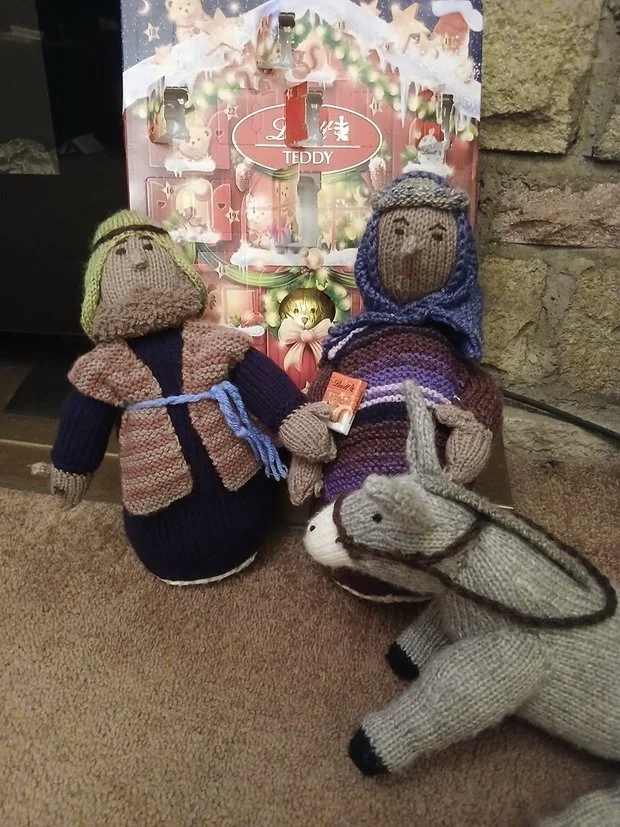 Two knitted dolls dressed in winter clothing, a girl with a yellow hat and blue staff, a woman with a purple shawl and headscarf, and a knitted donkey, are placed in front of an Advent calendar decorated for Christmas with a red and white theme, Christmas greenery, and a gold bell.