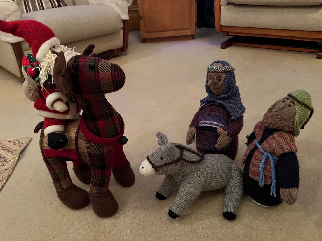 Christmas decorations with sock stuffed figures of a Christmas elf, two children, and a donkey on a beige carpeted floor, with couches and a wooden piece of furniture in the background.