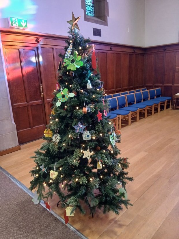 A decorated Christmas tree with a star topper, ornaments, and lights inside a room with wooden paneling and blue chairs along the wall.
