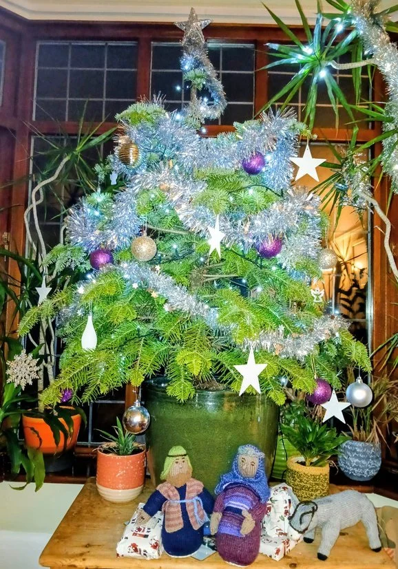 Christmas tree decorated with silver tinsel, purple, gold, and silver ornaments, and white stars. The tree is in a green pot on a wooden table, surrounded by holiday-themed plush figures and animal toys, and potted plants nearby.