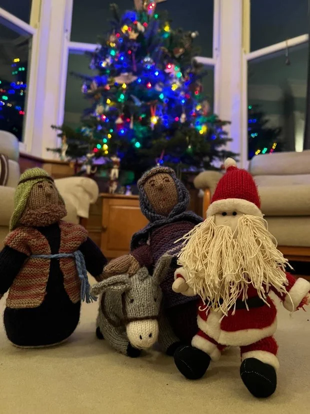 Three handmade Christmas dolls in front of a decorated Christmas tree with colorful lights inside a cozy living room.