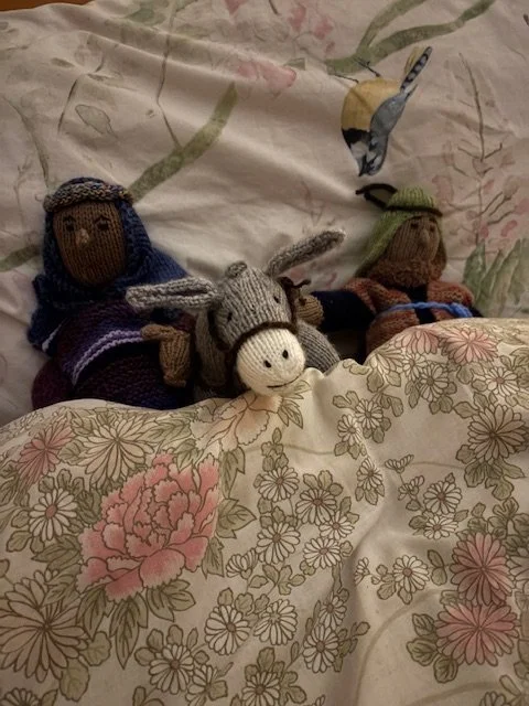 Three knitted stuffed animals, including a woman with a blue headscarf, a donkey, and a woman with a green hat, are emerging from a floral bedspread. A bird is flying overhead.