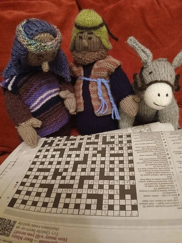 Three knitted stuffed animal toys sitting in front of a newspaper crossword puzzle on a red fabric background.