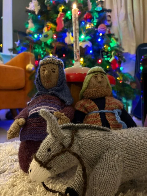 Knitted doll figures of Mary and Joseph with a plush donkey in front of a decorated Christmas tree.