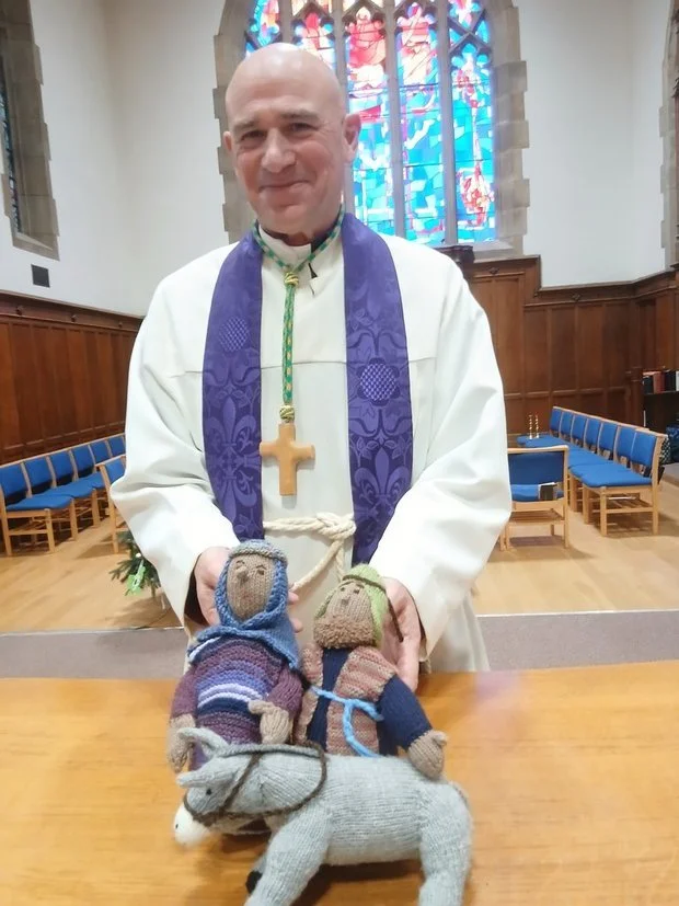A priest dressed in white robes with a purple stole and wooden cross necklace standing in a church with stained glass windows in the background. He is holding sock puppet figures, one of which is riding a stuffed animal, on a wooden table.