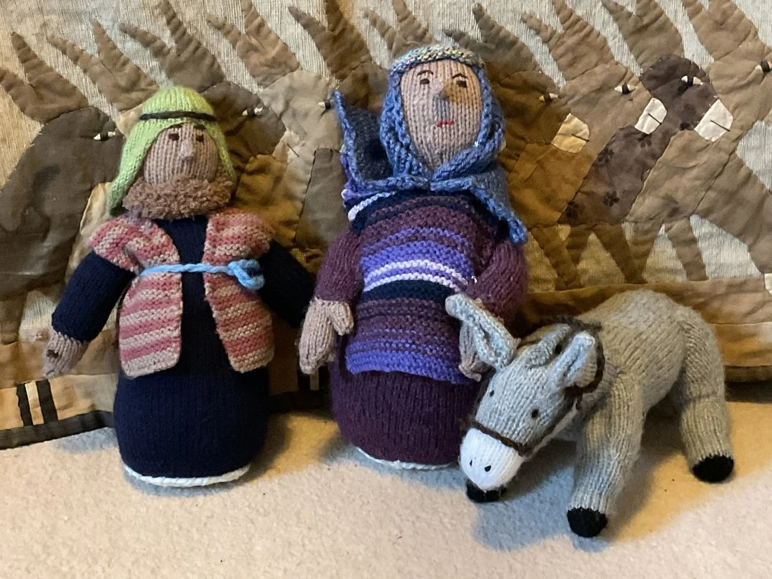 Two handmade knitted dolls and a stuffed toy horse positioned in front of a quilted wall hanging. The dolls are dressed in colorful knitted clothing and hats, with one holding a small blue rope. The quilted wall behind features animal patterns.