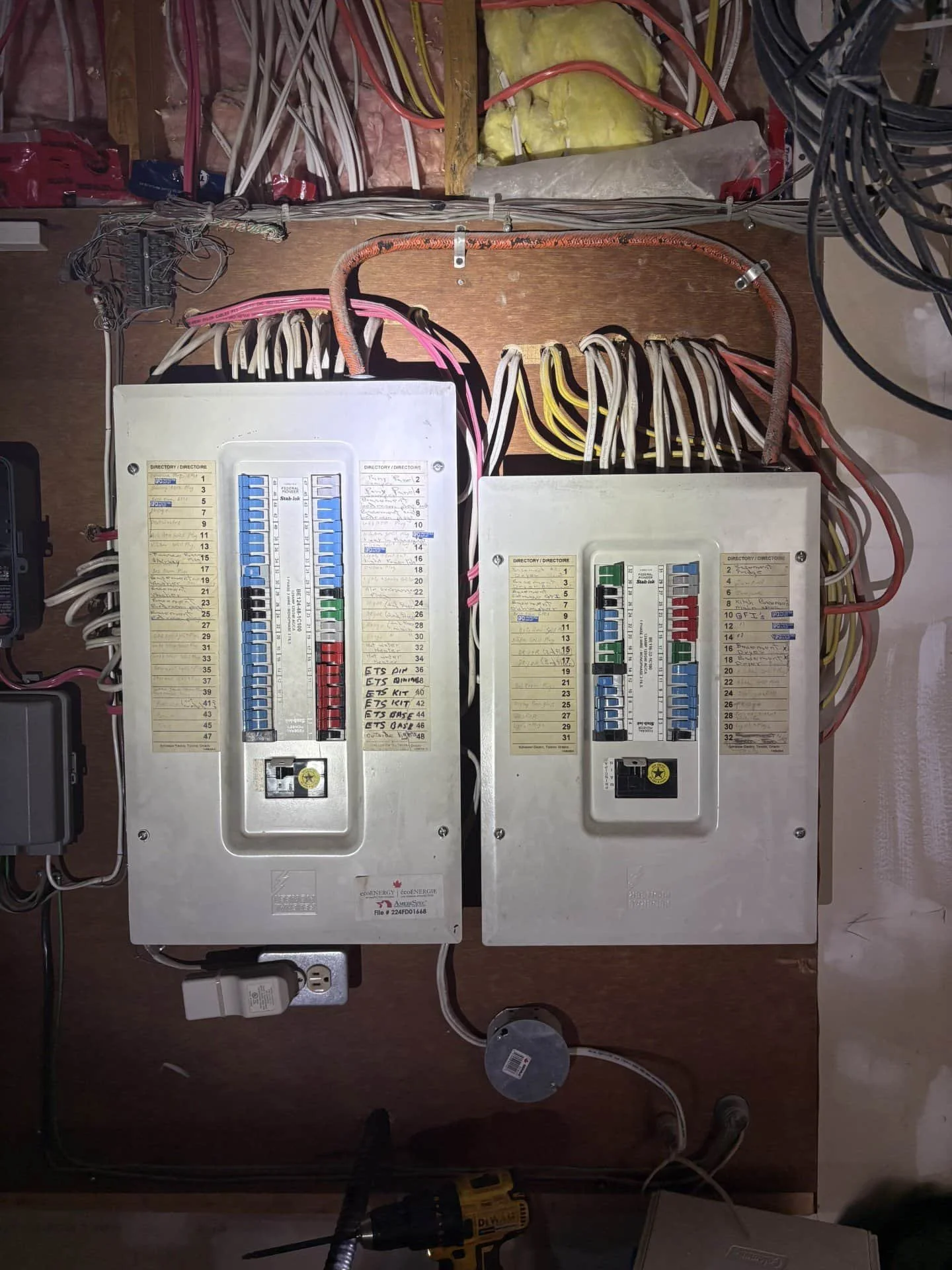 An old electrical panel with circuit breakers and wiring inside a wall. Two main breaker boxes are mounted on a wooden board with various colored wires connected to them.