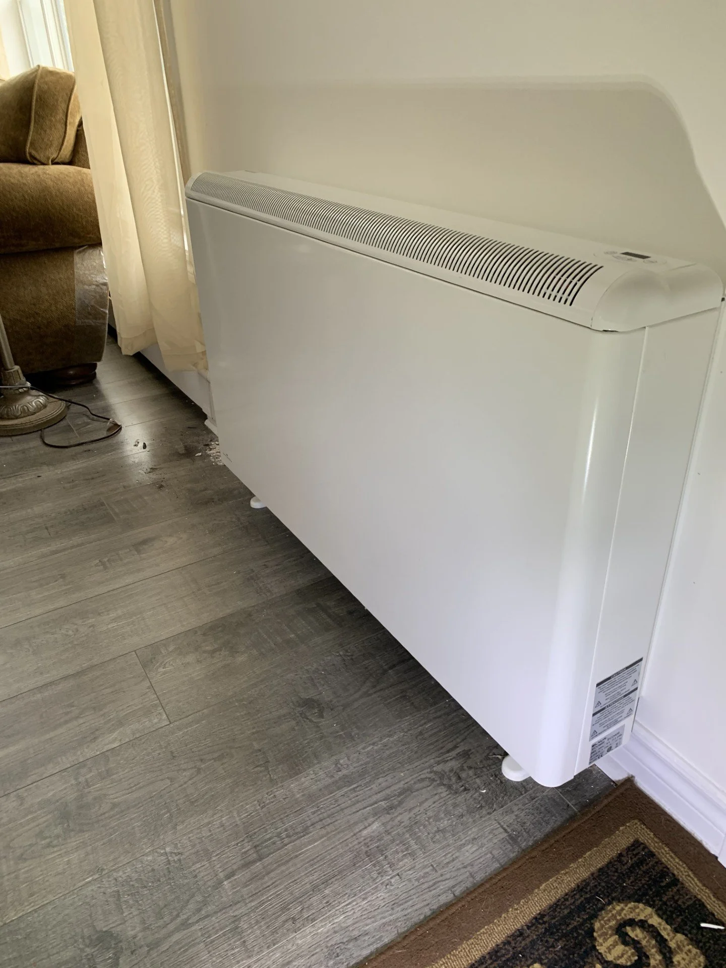 A white ETS heating system against a wall in a room with wood flooring and a patterned rug.