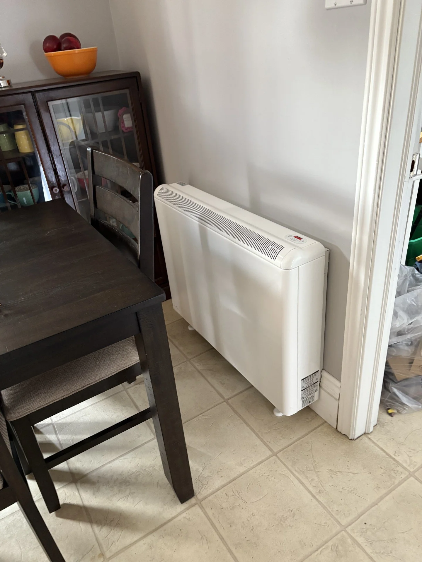 A white electric radiator heater placed on a beige tiled floor next to a dark wooden dining table and chair, with a wood china cabinet containing colorful dishes in the background.