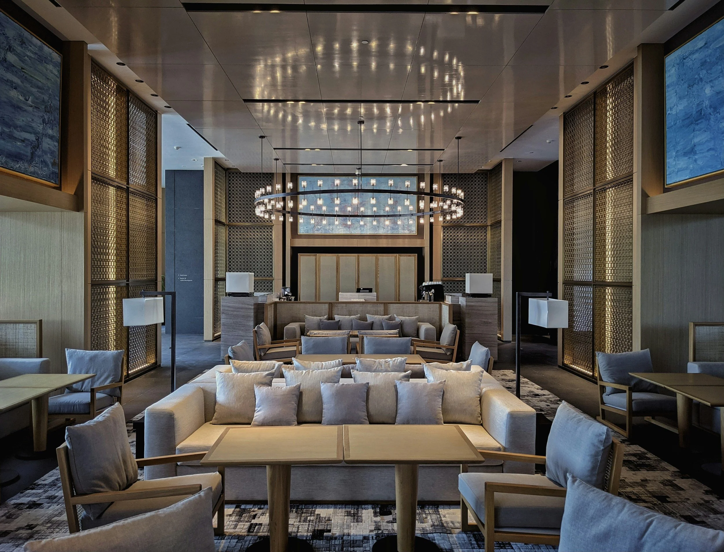Elegant hotel lobby with beige sofas, armchairs, and wooden tables, decorated with decorative pillows, illuminated by a large circular chandelier, with paintings and gold accents on the walls.