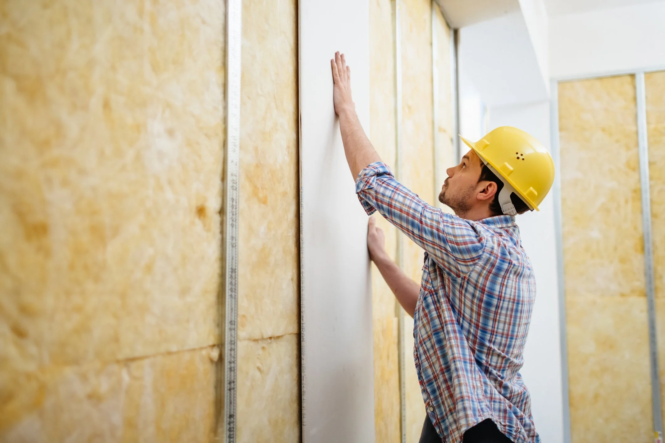 A construction worker in a yellow safety helmet and plaid shirt installs drywall on a wall with yellow insulation, using a measuring tool along the wall.