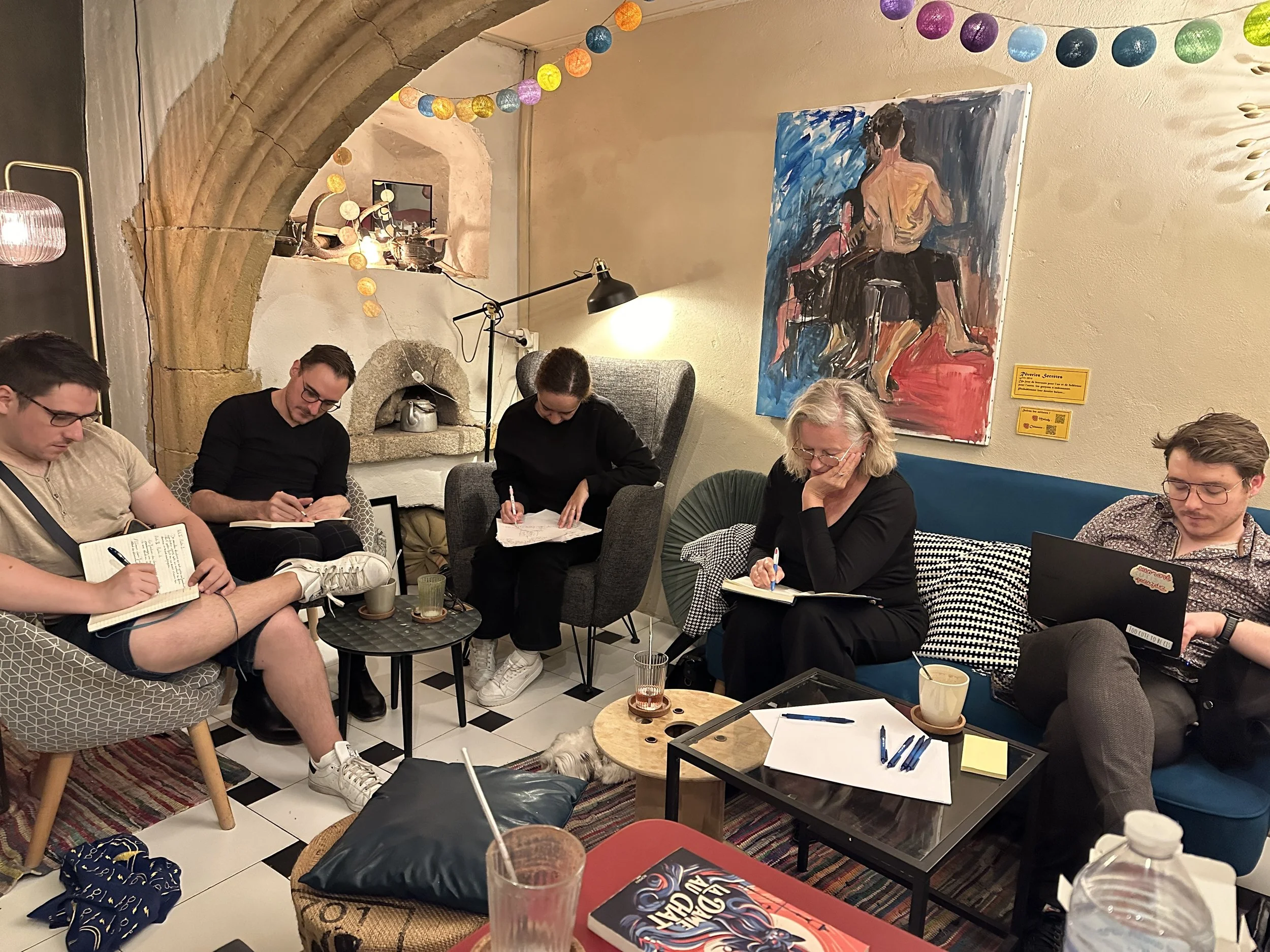 Un atelier d’écriture en cours : cinq personnes écrivent dans des fauteuils, autour de tables basses, dans la salle arrière d’un café