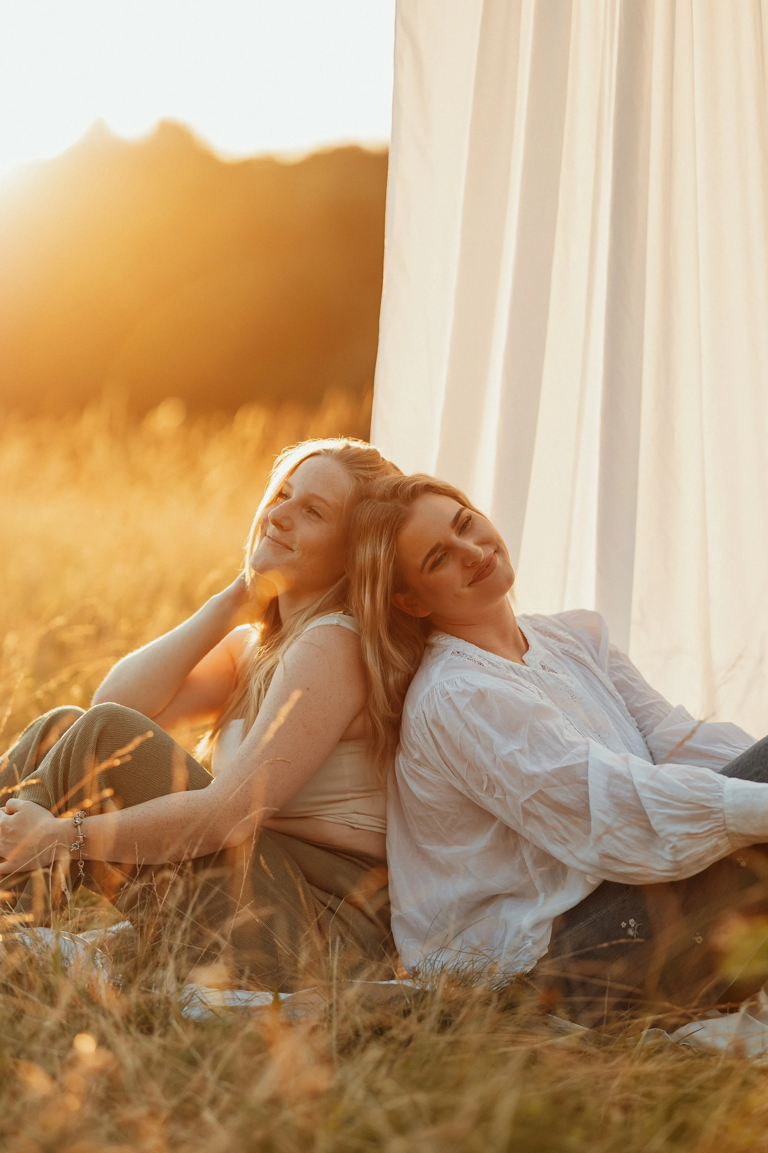 Zwei lachende Frauen sitzen im Feld an einem sonnigen Tag, mit weißen Kleidern und langen Haaren, vor einem weißen Vorhang im Freien.