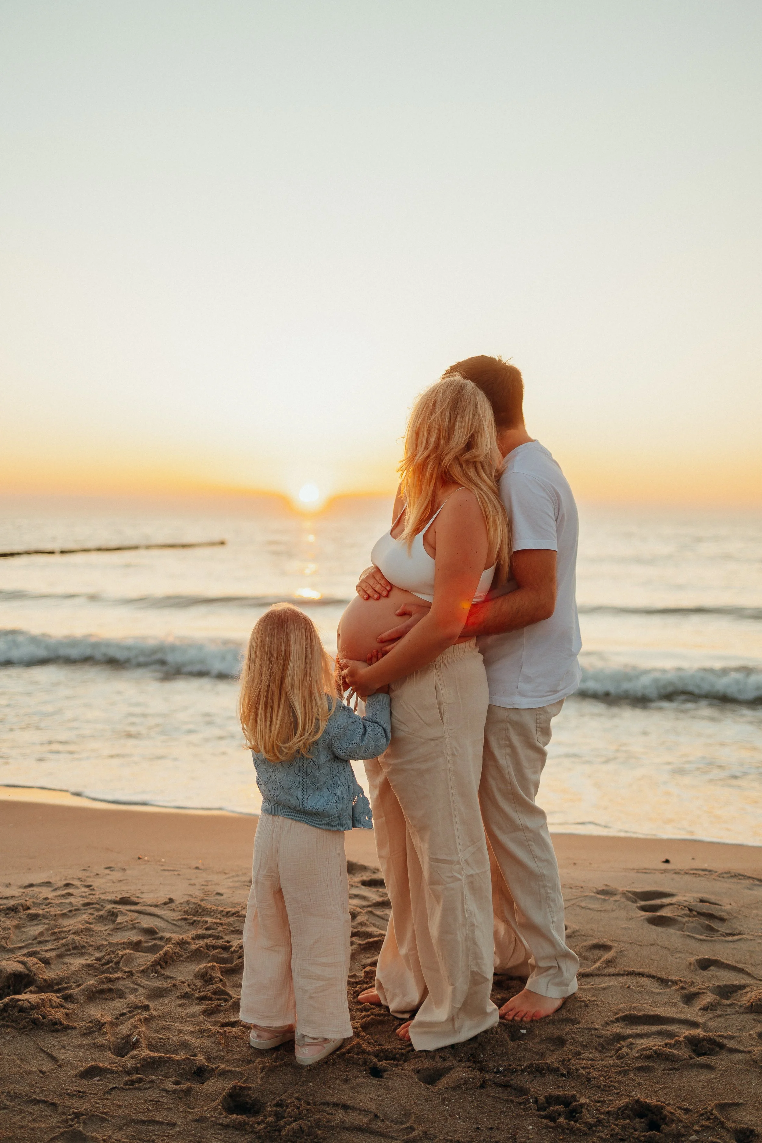 Eine Familie mit Mutter, Vater und einem kleinen Kind steht am Strand bei Sonnenuntergang, betrachtet das Baby.