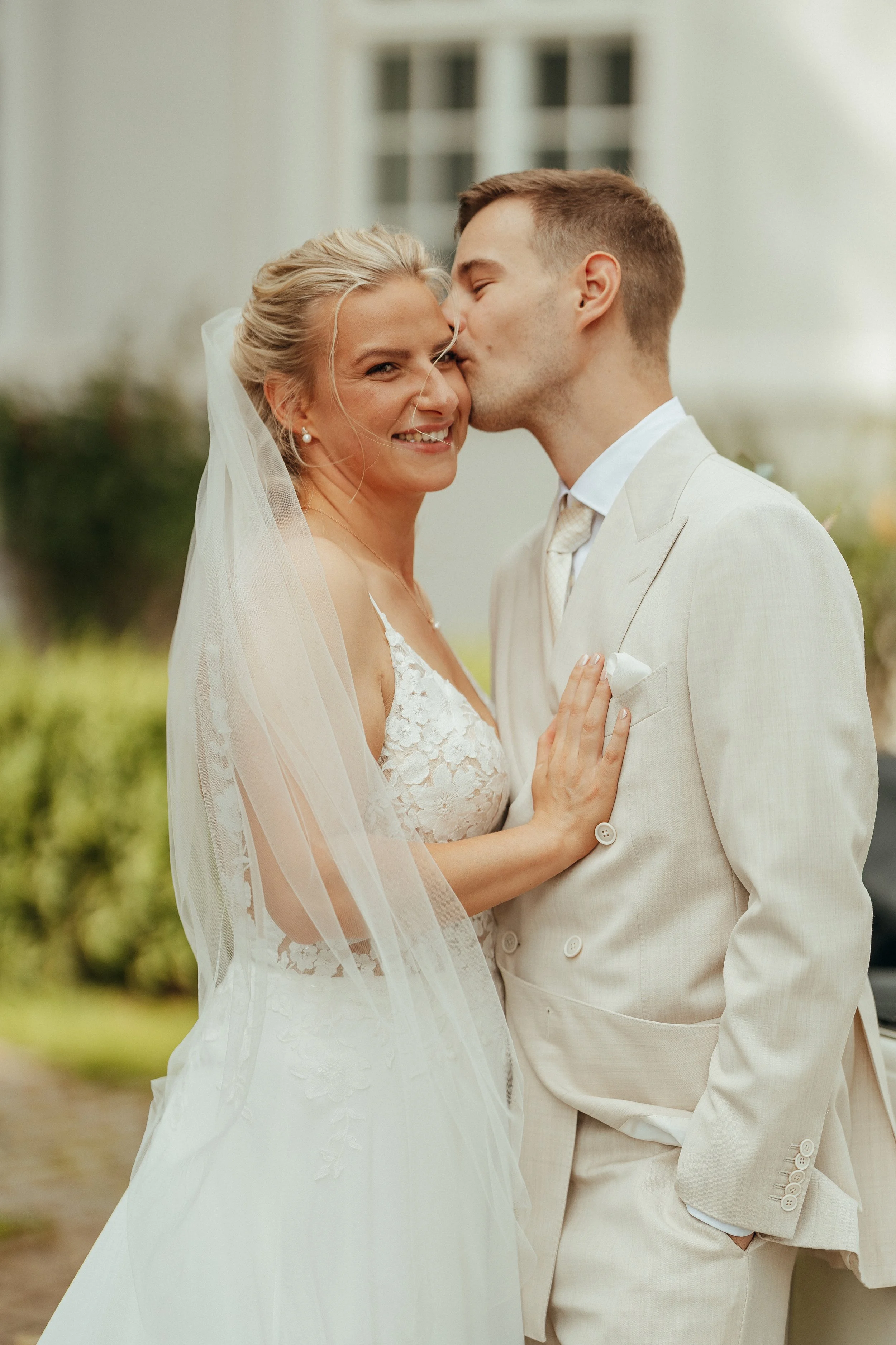 Ein Brautpaar, ein Mann und eine Frau, beim Küssen bei ihrer Hochzeit im Freien. Die Frau trägt ein weißes Hochzeitskleid und einen Schleier, der Mann trägt einen hellen Anzug.