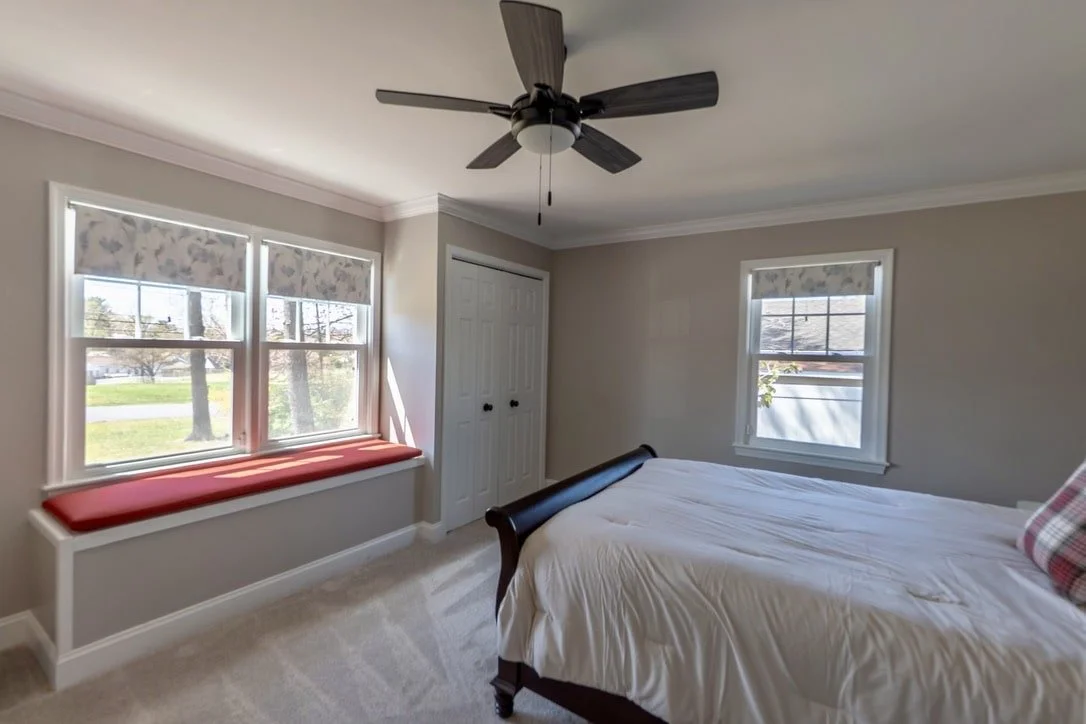 Bedroom with ceiling fan, white bedding, two windows with floral shades, red window seat cushion, and a closet with double doors.
