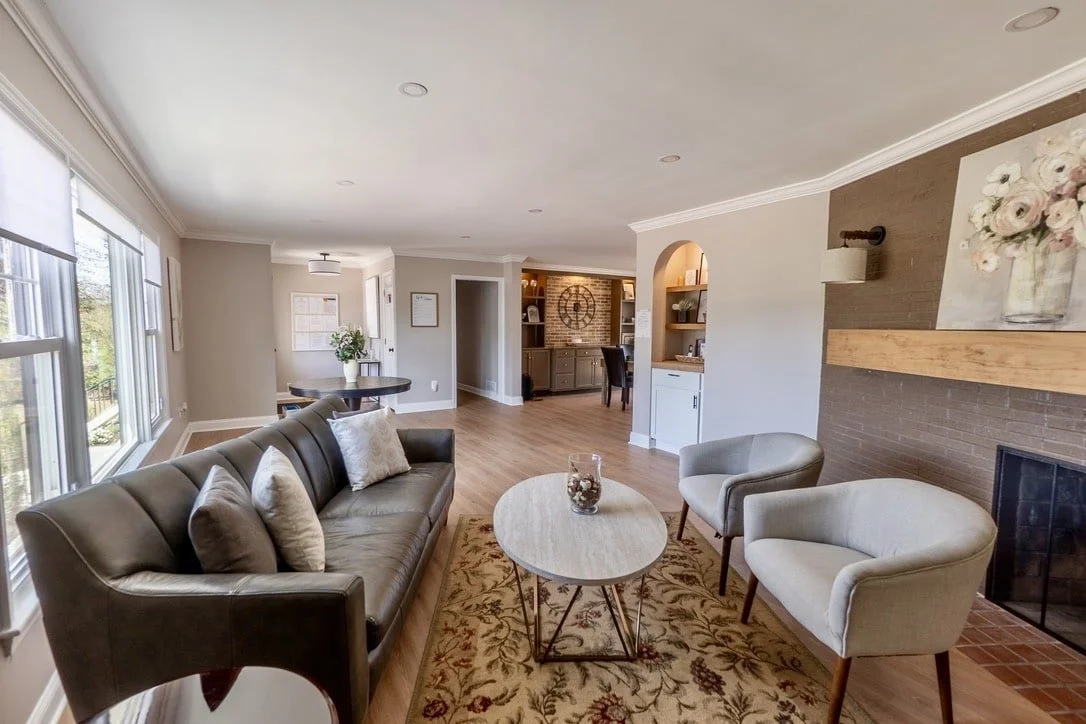 A modern living room with a gray sofa, two armchairs, a round coffee table, and a fireplace. The room features neutral colors, a patterned rug, a painting above the fireplace, and wooden floors. Large windows on the left allow natural light to enter.