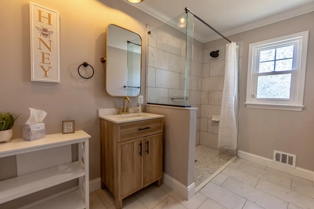 Modern bathroom with a wood vanity, rectangular mirror, and glass shower door. A "Honey" sign is on the wall, and a small shelf with tissues and a plant is visible.
