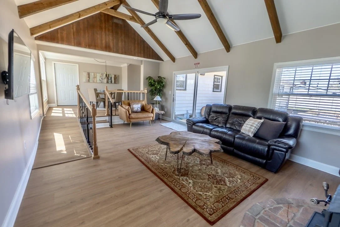 Modern living room with vaulted ceiling and exposed beams, featuring a black leather sofa, a wooden coffee table, a patterned rug, a leather armchair, a dining area in the background, and large windows providing natural light.