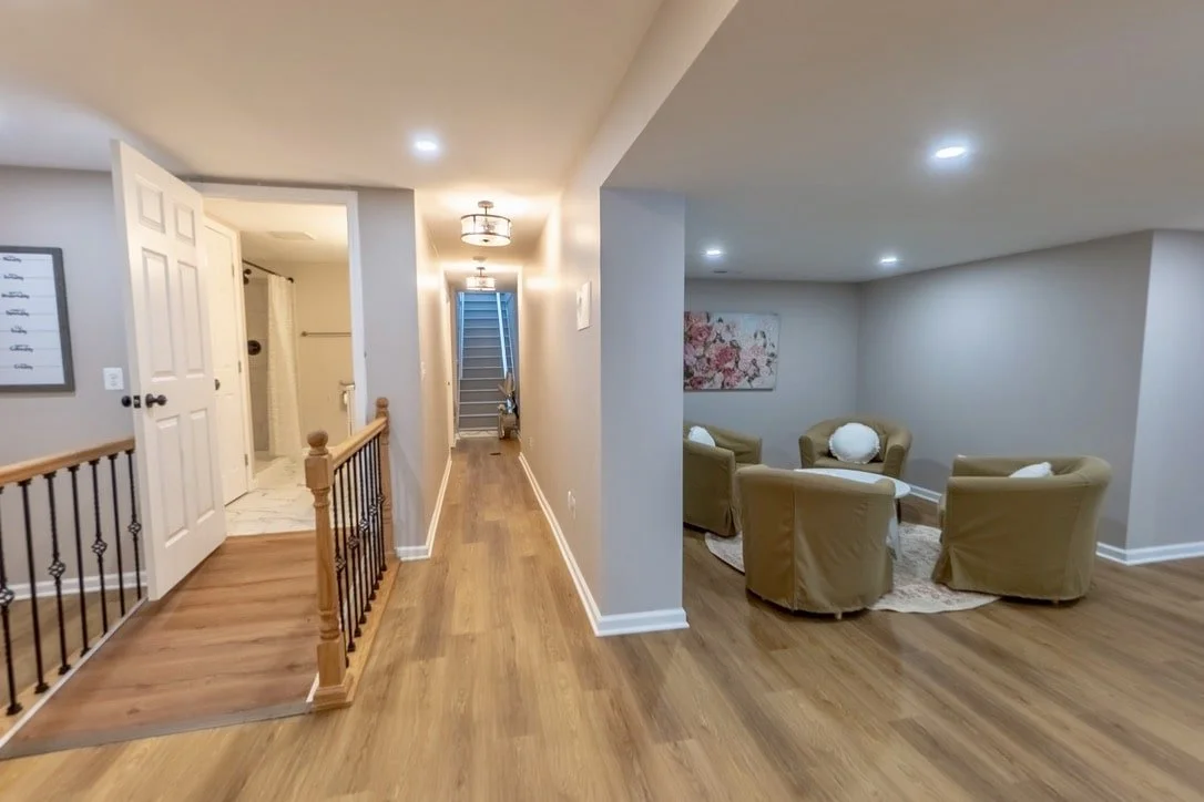 Basement interior with hallway, wooden floor, seating area with armchairs, round table, staircase, and open door to a bathroom.