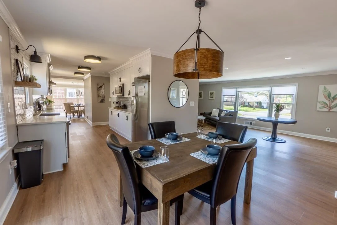 Modern open-concept kitchen and living area with wooden dining table, black leather chairs, contemporary lighting, and stainless steel appliances. Large windows provide natural light, while a circular mirror and decorative plants enhance the decor.
