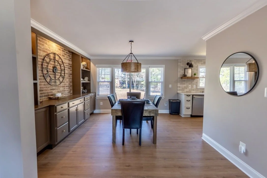 Modern dining area with wooden table and chairs, brick wall, large wall clock, built-in cabinets, pendant light, and a round mirror.