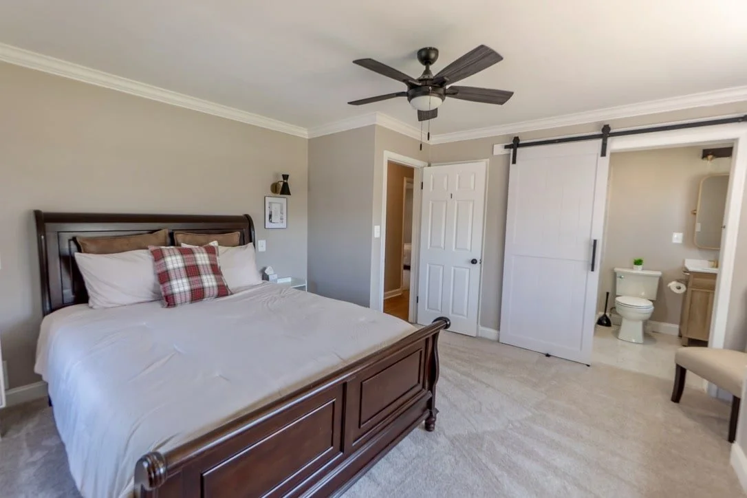 Modern bedroom with a large bed, decorative pillows, ceiling fan, and barn door leading to a bathroom.