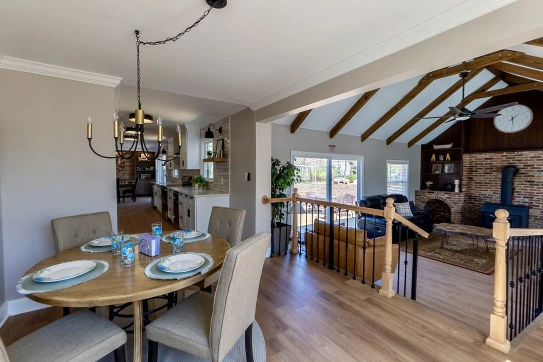 Open-concept living and dining area with round dining table, chandelier, kitchen with white cabinets, living room with exposed wooden beams, brick fireplace, and glass doors to outside.