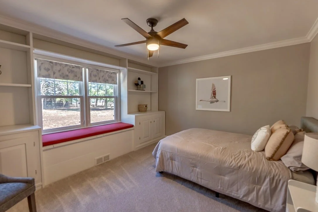 Small bedroom with neutral decor, featuring a bed with beige bedding, a window seat with a red cushion, and built-in shelving. A ceiling fan is above, and a framed sailing artwork hangs on the wall.