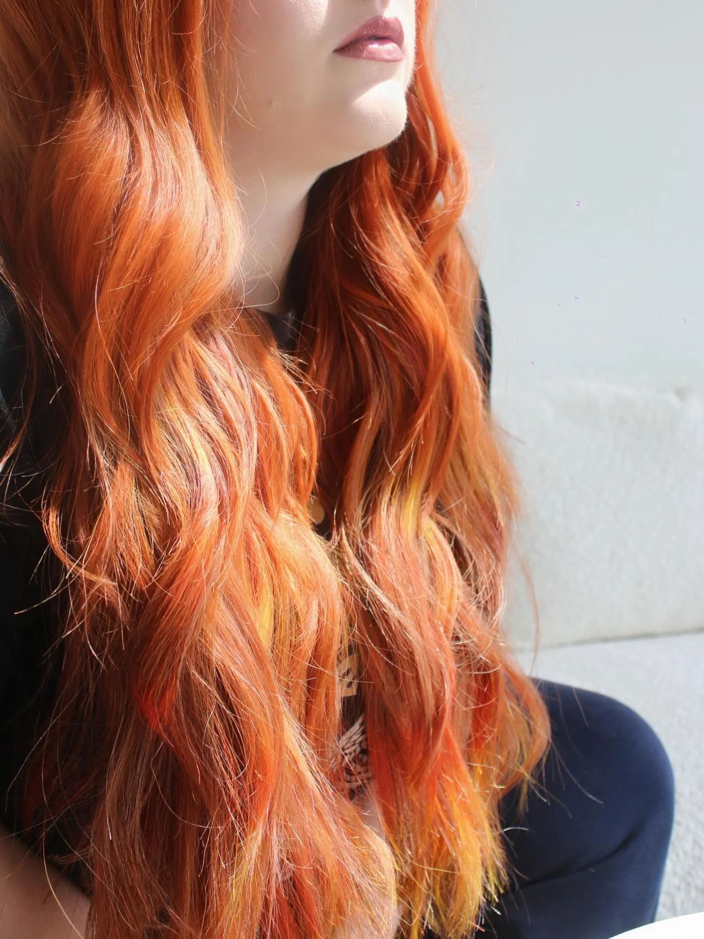 Close-up of a woman with long, wavy, orange-red hair partially covering her face and lips, sitting against a light-colored wall.