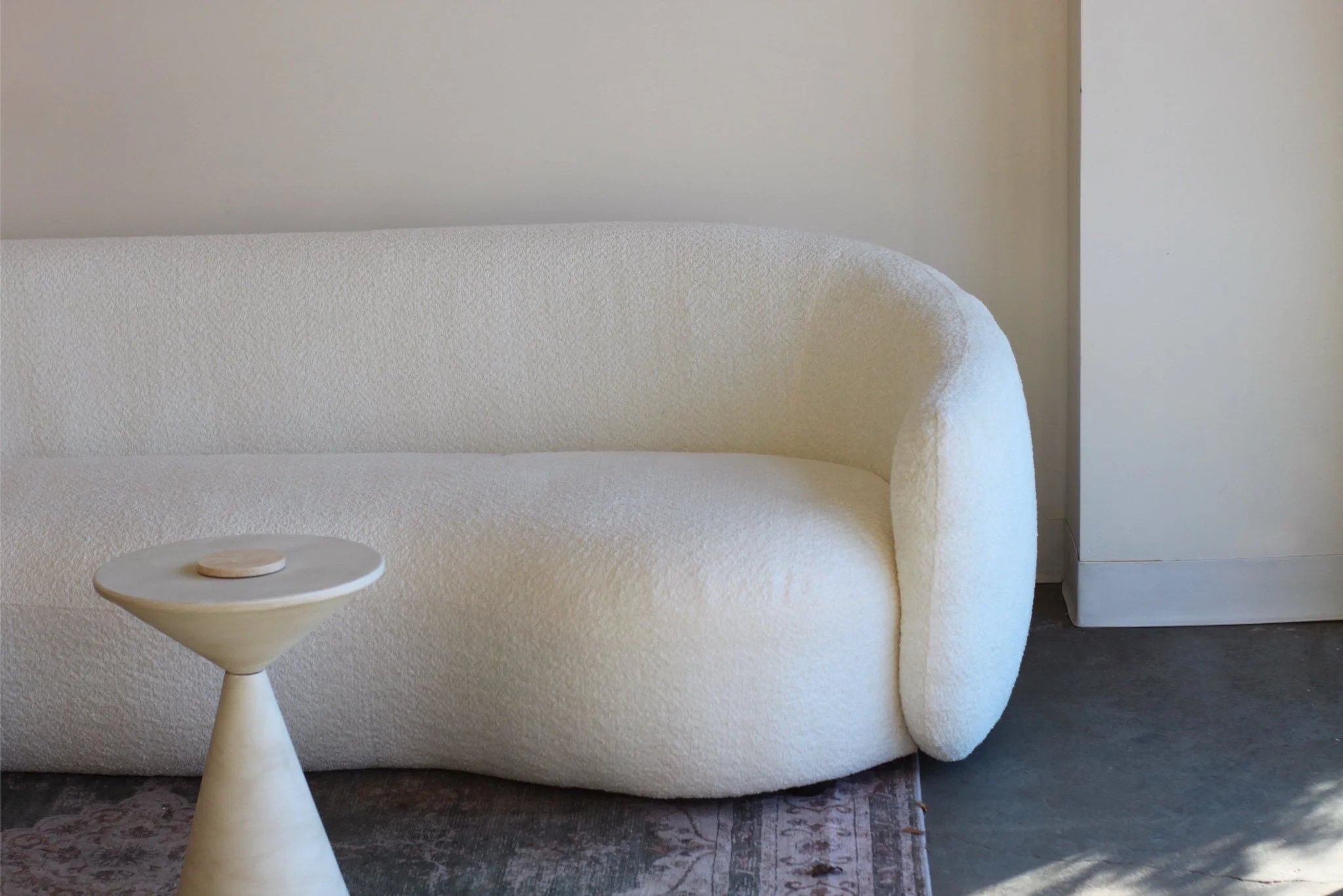 A white textured armchair with rounded armrests next to a small round side table with a wooden top, set against a plain wall and partially on a dark-patterned rug.