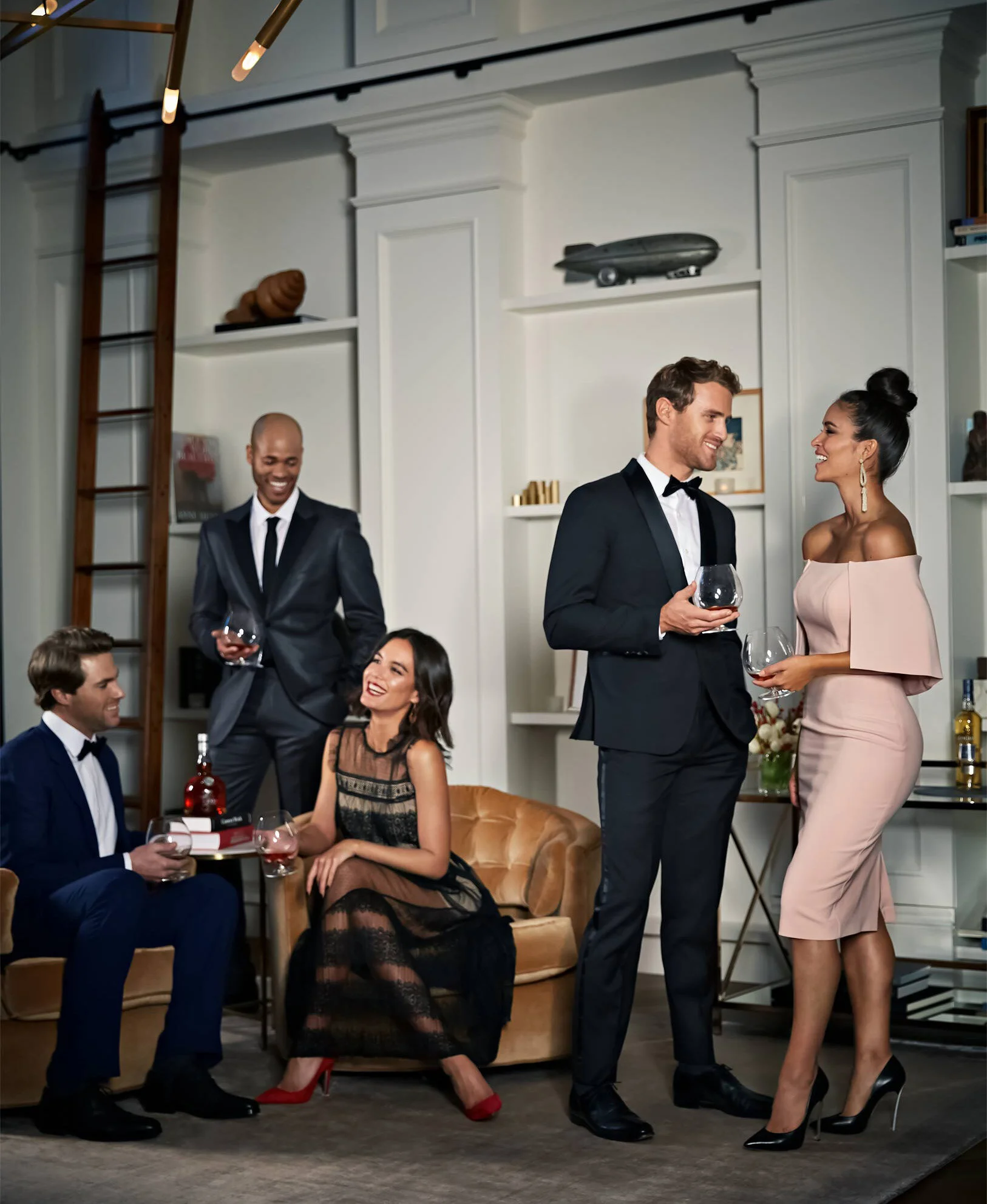 Group of five adults at a formal party in a modern living room, with two couples chatting, one man standing and talking to a woman, two people sitting on a tan couch enjoying drinks, and one man standing in the background next to a bookshelf.