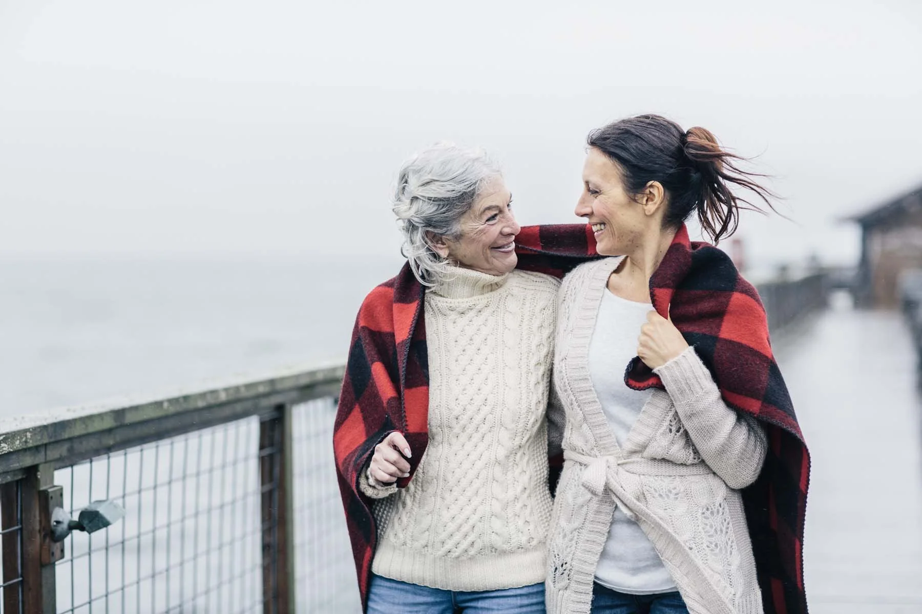 Two women, an elderly woman and a middle-aged woman, smiling and embracing each other outdoors by the water on a cloudy day, wrapped in a red and black plaid blanket.