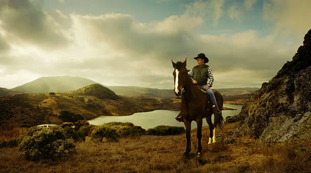 A woman riding a horse in a scenic landscape with hills, a lake, and cloudy sky.