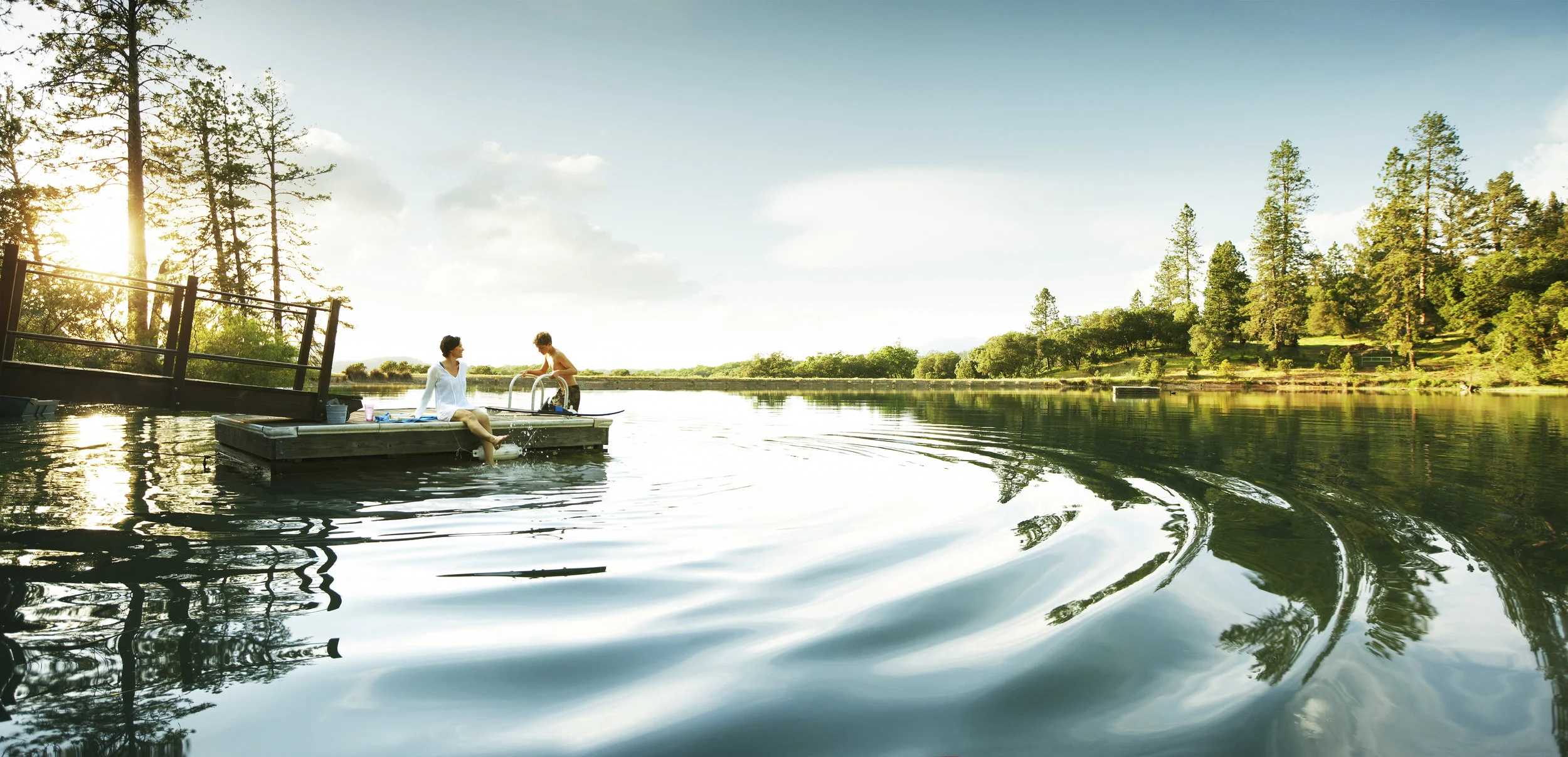 Two kids and an adult on a wooden dock by a lake with trees in the background during sunset.