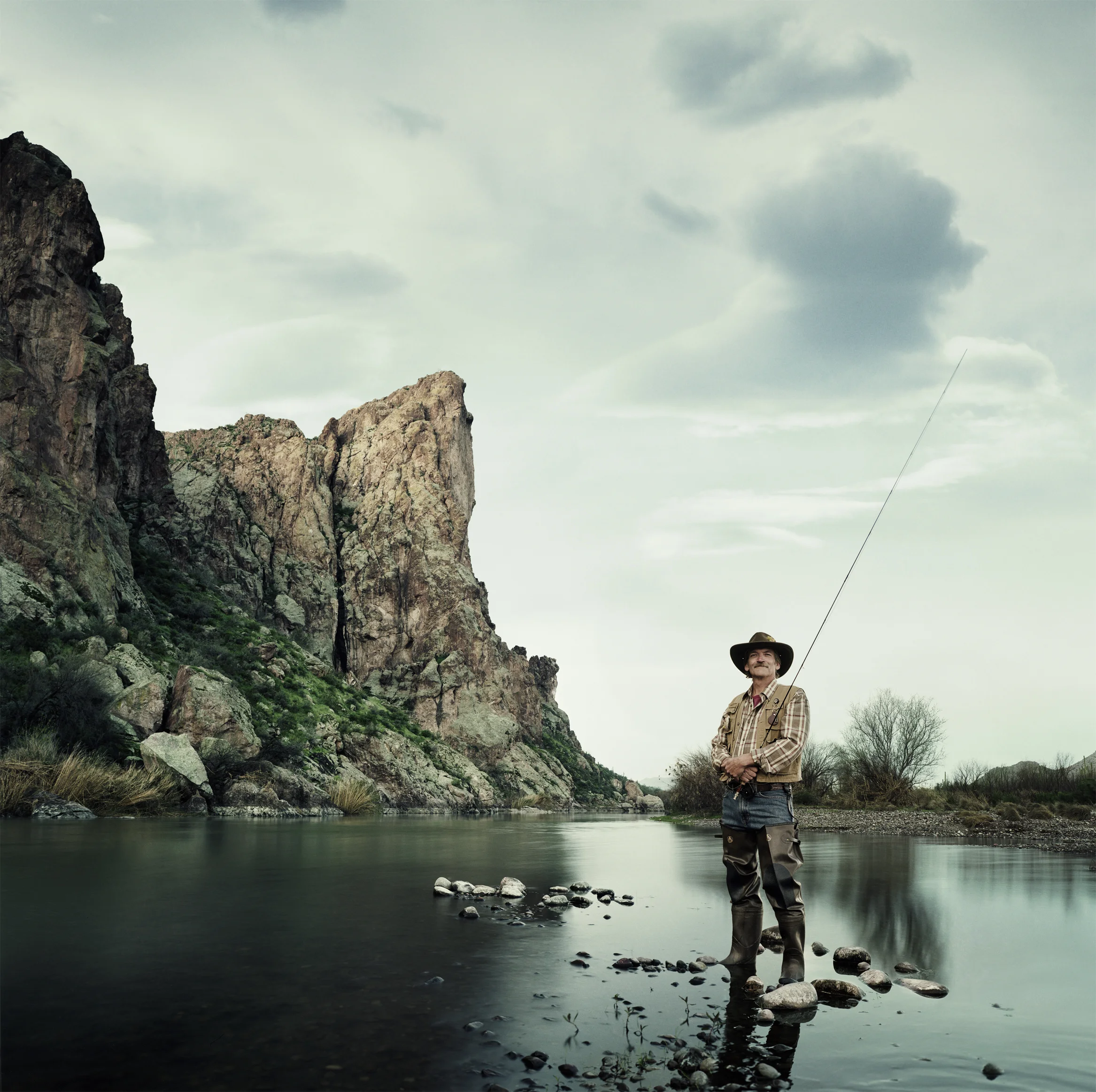 A man standing in a river with a fishing rod, surrounded by rocky cliffs and sparse trees under a cloudy sky.