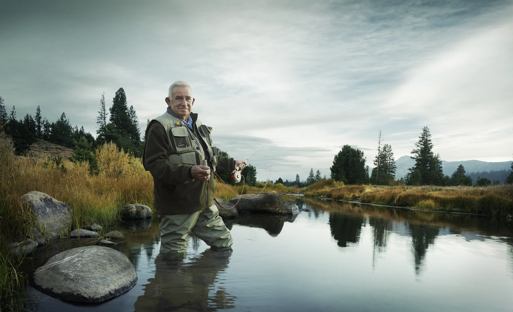 Older man fishing in a creek with trees and mountains in the background.