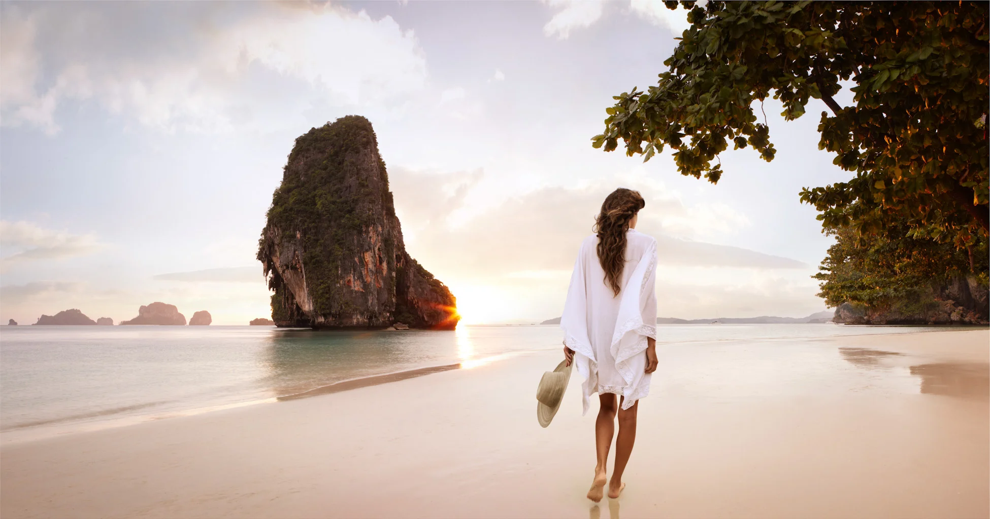 A woman in a white dress walking barefoot on a beach at sunset, holding a wide-brimmed hat, with limestone islands in the background and a tree on the right.