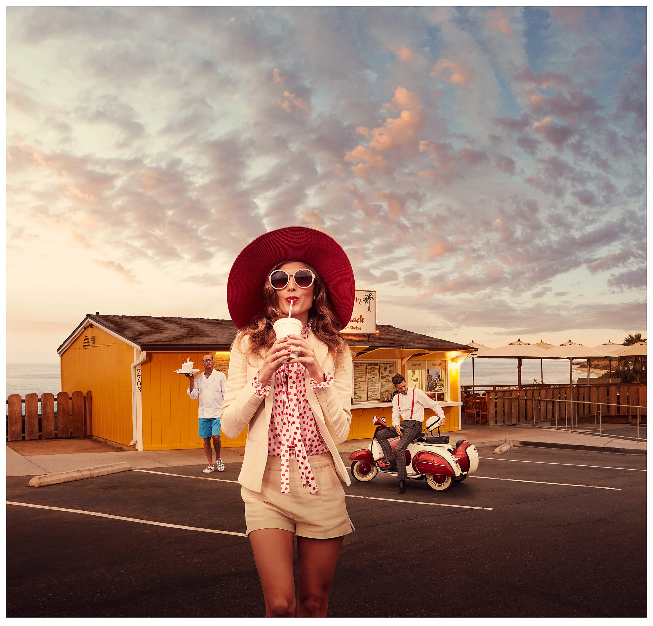 A woman wearing a red wide-brim hat, large sunglasses, a cream blazer, and cream shorts drinks from a cup with a straw in a parking lot near a yellow beachside shack at sunset. Two men are in the background, one holding a tray and the other sitting o