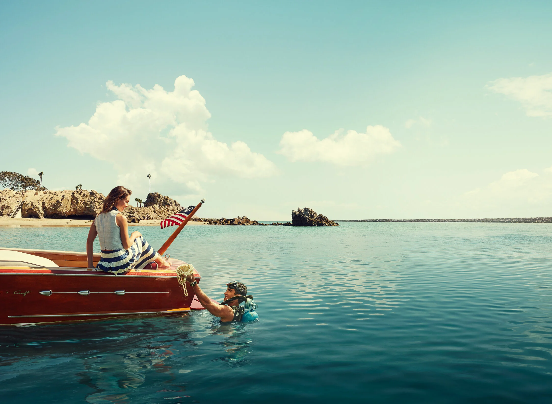 A woman sitting on a boat holding an American flag, with a diver in the water reaching up to her, near a rocky shoreline under a partly cloudy sky.
