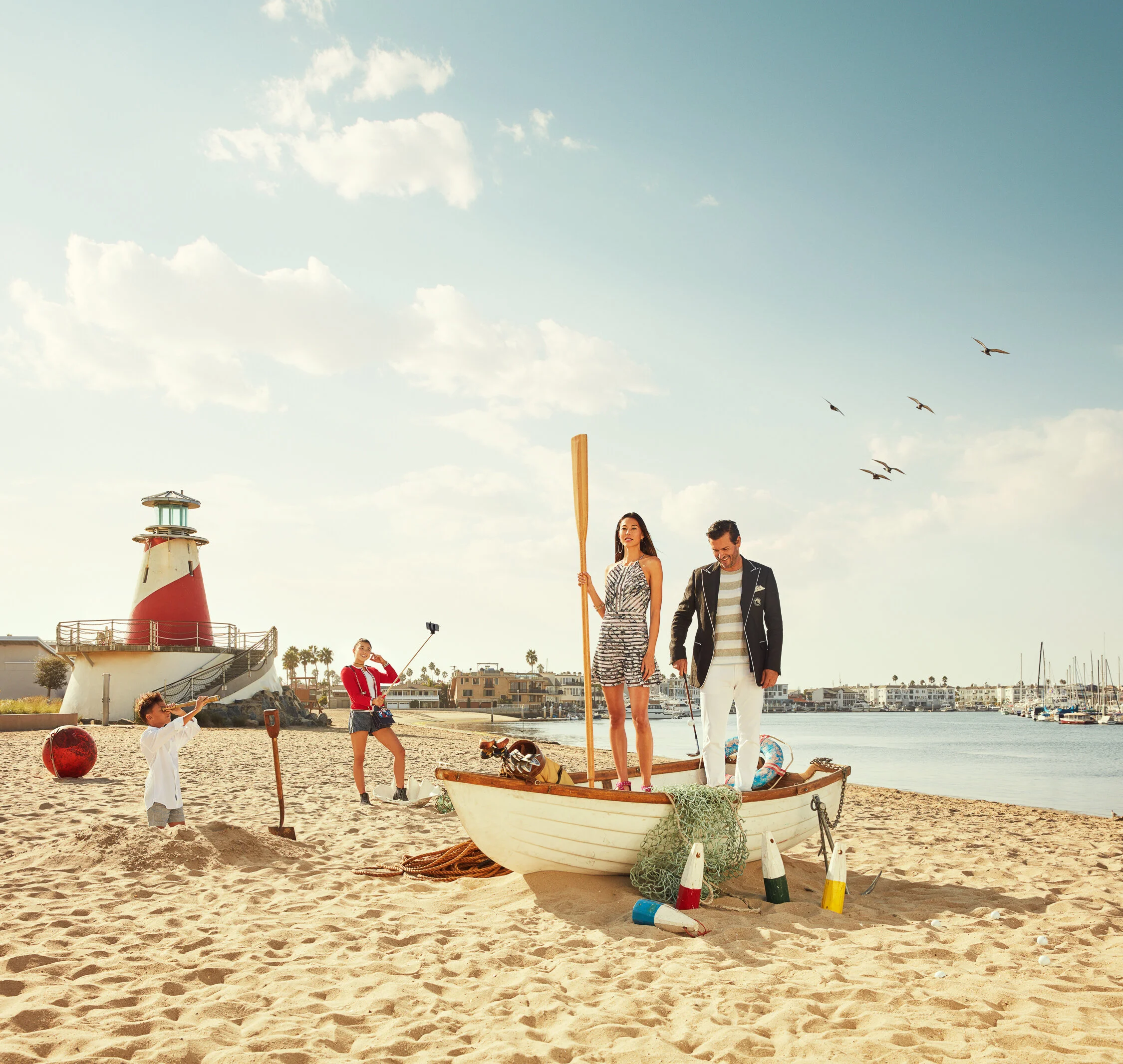 A family on a beach with a small boat, children playing in sand, lighthouse in the background, and a marina with boats docked along the water, under a partly cloudy sky.