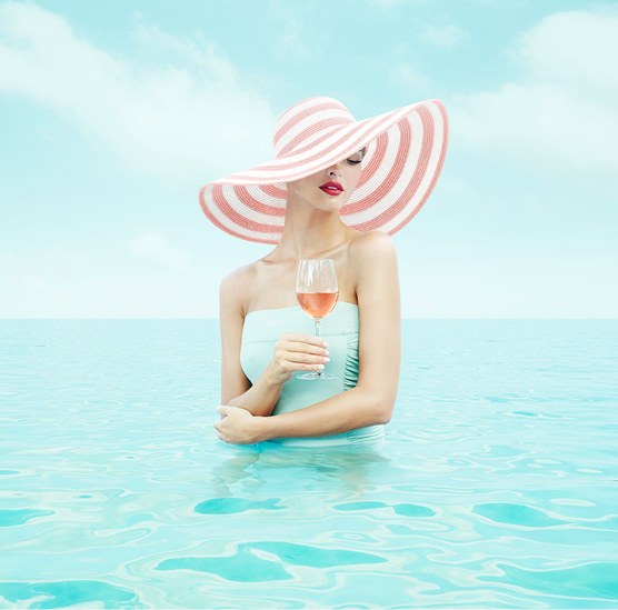 Woman in water wearing a large striped sunhat, holding a glass of rosé wine, against a bright sky and calm water.