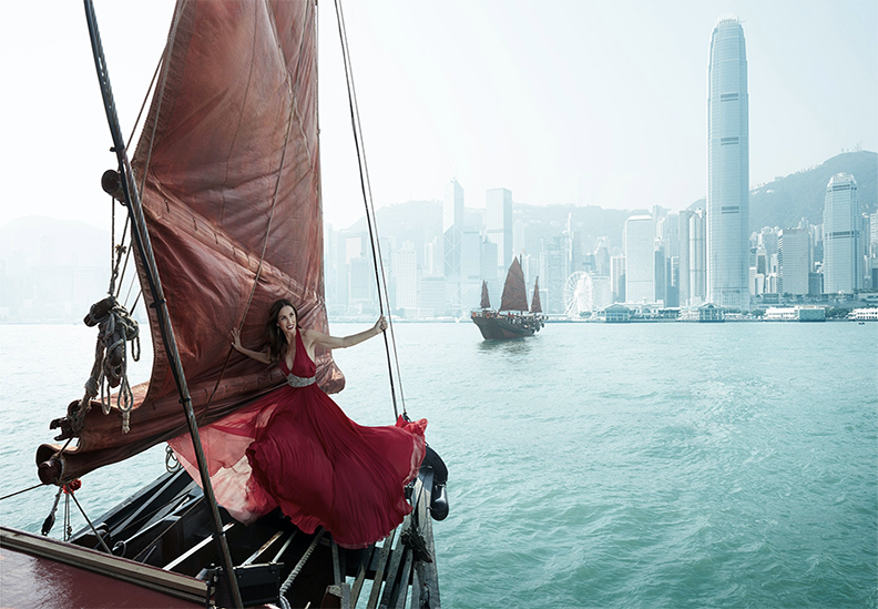 A woman in a red dress sitting on a sailboat with a city skyline in the background, including tall skyscrapers and a traditional junk boat in the water.
