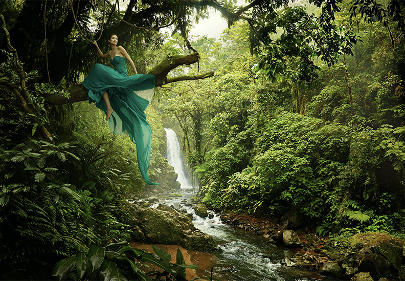 A woman in a flowing turquoise dress sitting on a tree branch in a lush, green jungle by a waterfall and flowing stream.