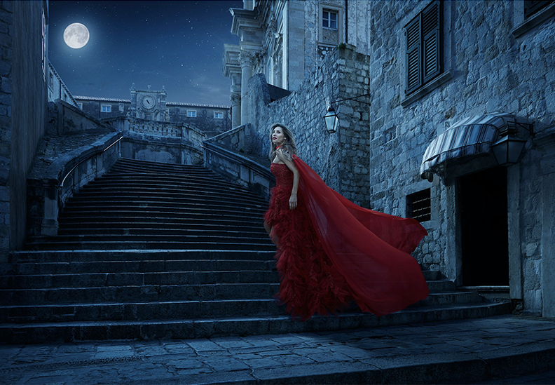 A woman in a red gown with a long train stands on stone stairs outside a historic stone building at night, illuminated by moonlight and street lamps.