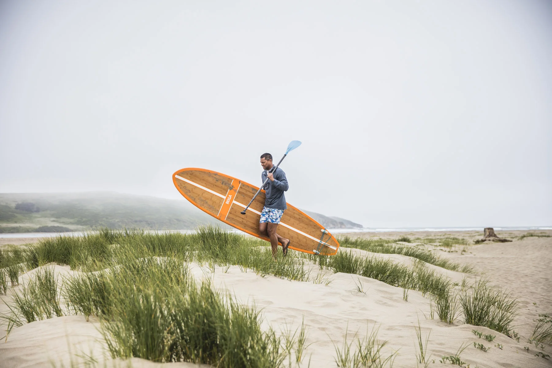 A man holding a paddle and standing on a surfboard on a sandy beach with grassy dunes and an overcast sky.