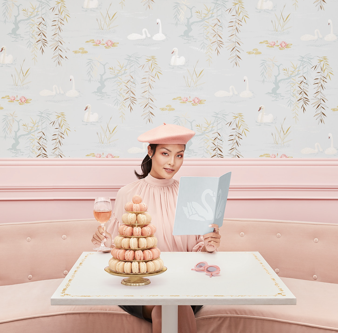 A woman dressed in pink with a pink beret, sitting at a pink booth in a pastel-colored swan-themed decor, holding a menu in one hand and a glass of rosé wine in the other, with a tower of macarons on the table.