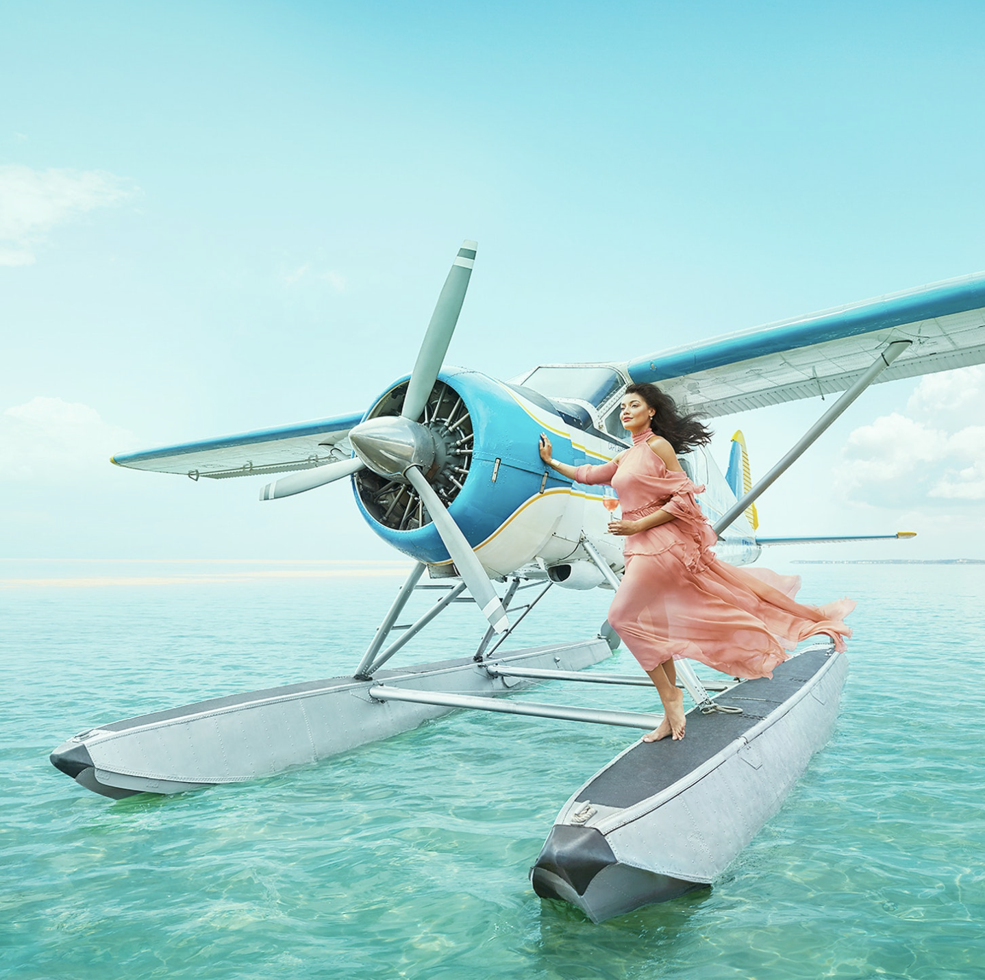 Woman in a pink flowing dress standing on a floatation device next to a blue and white seaplane on calm ocean water, with a partly cloudy sky.